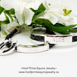 May include: Silver bracelets and a pendant with black woven accents. The bracelets and pendant have the name "KLOE" engraved on them. White flowers and green leaves are in the background. The jewelry is from Hoof Prints Equine Jewelry.