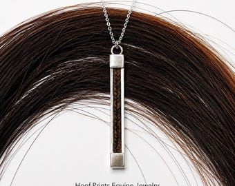 May include: A silver-toned vertical bar pendant necklace featuring a central inlay of braided dark brown hair. The pendant hangs from a delicate silver chain. Text at the bottom reads: Hoof Prints Equine Jewelry www.hoofprintsequinejewelry.ca