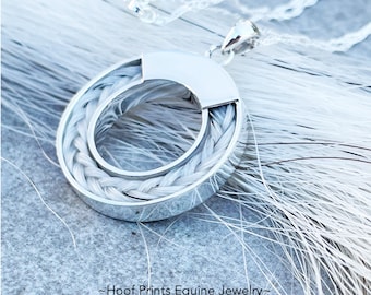 May include: A silver pendant necklace with a braided, oval-shaped design. The pendant features a polished silver frame with a woven, light gray material inside. The necklace is displayed on a light gray surface with white strands.