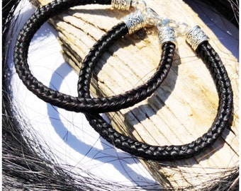 May include: Two black braided horsehair bracelets with silver decorative clasps. The bracelets are on a wooden surface.