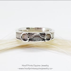 May include: A silver ring with a horsehair inlay and a silver snaffle bit design. The ring is on a white horsehair background.
