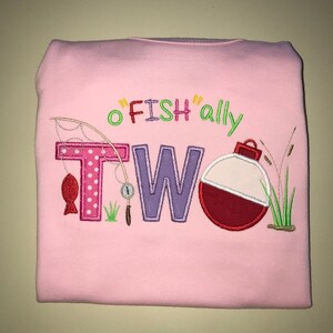 May include: Pink toddler shirt with the text "o'fish'ally two" embroidered in colorful letters. The design includes a fishing rod, a fish, a bobber, and grass.