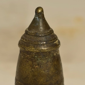 Puede incluir: Un pequeño tapón de botella antiguo de latón con una parte superior cónica y un acabado desgastado y envejecido. El tapón tiene un diseño decorativo alrededor de la parte superior e inferior.