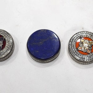 May include: Three round decorative boxes with silver-colored metal rims. Two have ornate designs with red and orange backgrounds, and the third is a solid blue. The boxes are likely for jewelry or small items.
