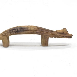 May include: A light brown, carved wooden drawer pull shaped like an alligator. The handle features a textured, scaled back, a defined head with snout, and a tail, supported by two vertical legs that extend downward for mounting purposes.