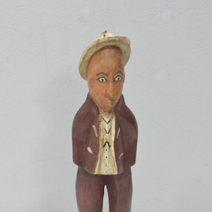 May include: A wooden figurine of a person wearing a brown jacket, white shirt, and a white hat. The figurine is standing on a wooden base.