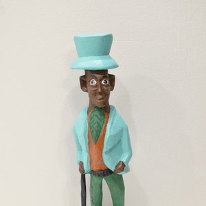 May include: A wooden figurine of a person wearing a blue hat, a blue jacket, an orange shirt, green pants, and holding a black walking stick. The figurine is standing on a green base.