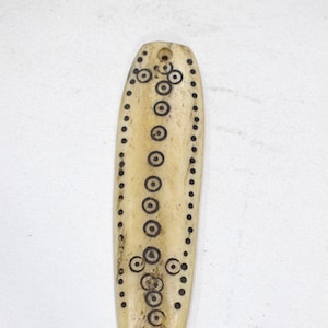 May include: A cream-coloured, elongated pendant with a hole at the top. The pendant is decorated with black circular and dot patterns. The design includes a central line of circles and dotted edges.