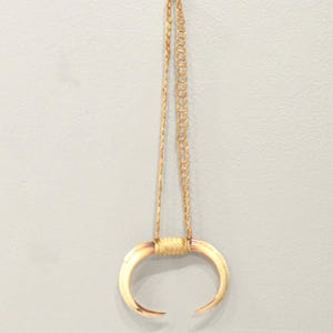 May include: A pendant necklace featuring a curved, off-white horn-shaped pendant. The pendant is suspended from a gold-colored chain and cord. The necklace is displayed against a light gray background.