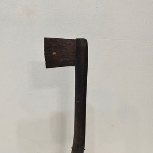 May include: A vintage axe with a dark wooden handle and a dark, rectangular blade. The axe is mounted on a black metal stand, showcasing its rustic design. The axe is a dark brown color.