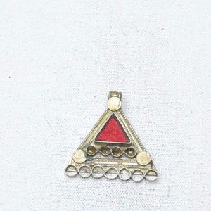 Puede incluir: Un colgante de triángulo de plata con un centro rojo y detalles decorativos. El colgante tiene un bucle en la parte superior para sujetarlo a una cadena.