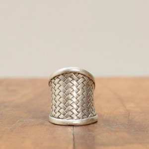 May include: A silver ring with a woven design. The ring is wide and has a rounded top.