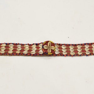 Puede incluir: Un cinturón rojo de cuentas con un diseño de conchas blancas y una hebilla circular de concha con un patrón de flor amarilla y blanca.