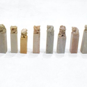 May include: A collection of rectangular stone seals, each topped with a carved animal figure. The seals vary in color, including beige, gray, and pink. The animal carvings feature detailed designs, showcasing a variety of creatures.