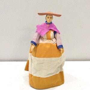 May include: A handmade doll wearing a traditional outfit with a wide-brimmed hat, a pink shawl, a white and orange dress, and a yellow skirt with white stripes.