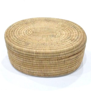 Puede incluir: Una cesta de almacenamiento ovalada tejida de forma natural con tapa. La cesta está hecha de ratán marrón claro y tiene un acabado natural.
