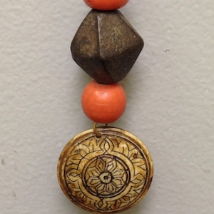 May include: A handcrafted necklace featuring a brown cord, orange beads, a dark geometric bead, and a carved circular pendant with an intricate floral design. The pendant is a light tan color.