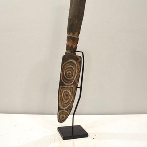 May include: A wooden carving with a long handle and a blade decorated with concentric circles and geometric patterns. The carving is mounted on a black metal stand.