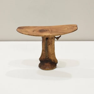 May include: A wooden stool with a curved top and a single, tall, cylindrical leg. The stool is made of a light brown wood and has a natural finish.