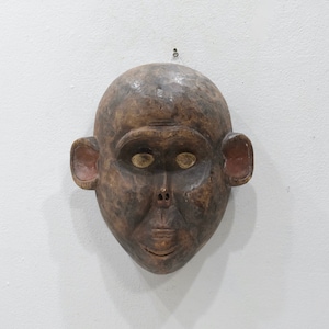 May include: A wooden mask depicting a stylized human face with large eyes and a wide mouth. The mask is painted in shades of brown and has a reddish-brown hue around the ears.