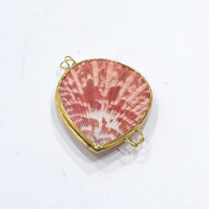 May include: A heart-shaped pendant featuring a natural seashell with a reddish-pink and white radial pattern, encased in a thin gold-toned metal frame with two small wire loops attached to the sides for hanging or connecting to a chain.
