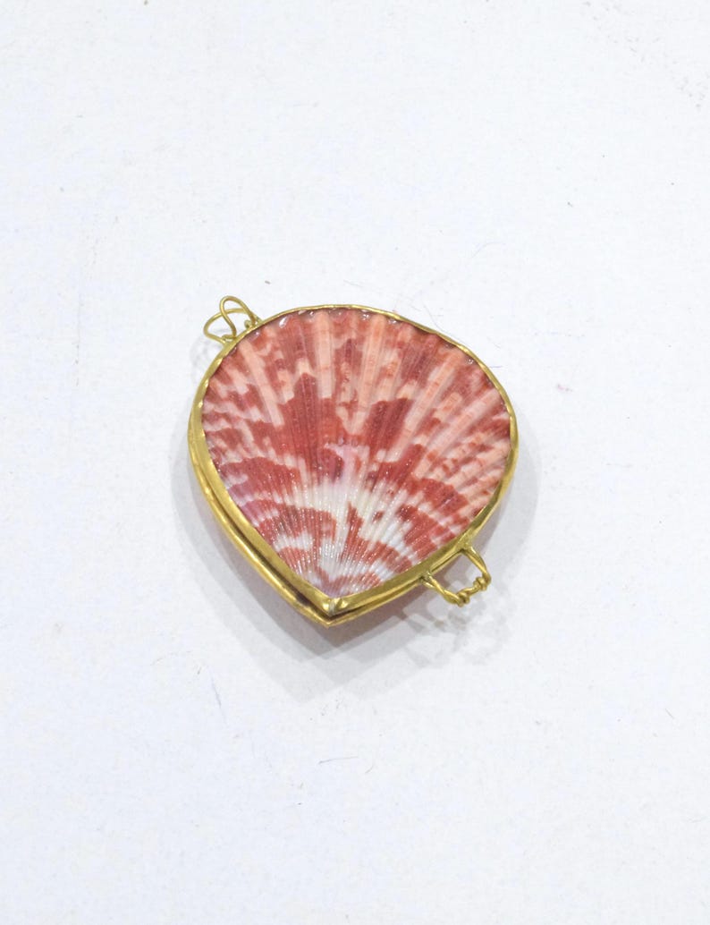 May include: A heart-shaped pendant featuring a natural seashell with a reddish-pink and white radial pattern, encased in a thin gold-toned metal frame with two small wire loops attached to the sides for hanging or connecting to a chain.