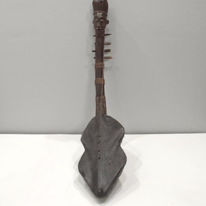 May include: A dark brown, handcrafted stringed instrument with a carved face at the top. The body has a unique, asymmetrical shape. The instrument appears to be made of wood and has several small pegs along the neck.