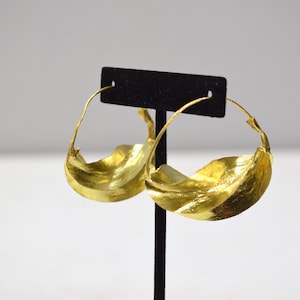 May include: A pair of gold hoop earrings with a wavy, organic design. The earrings are displayed on a black stand.