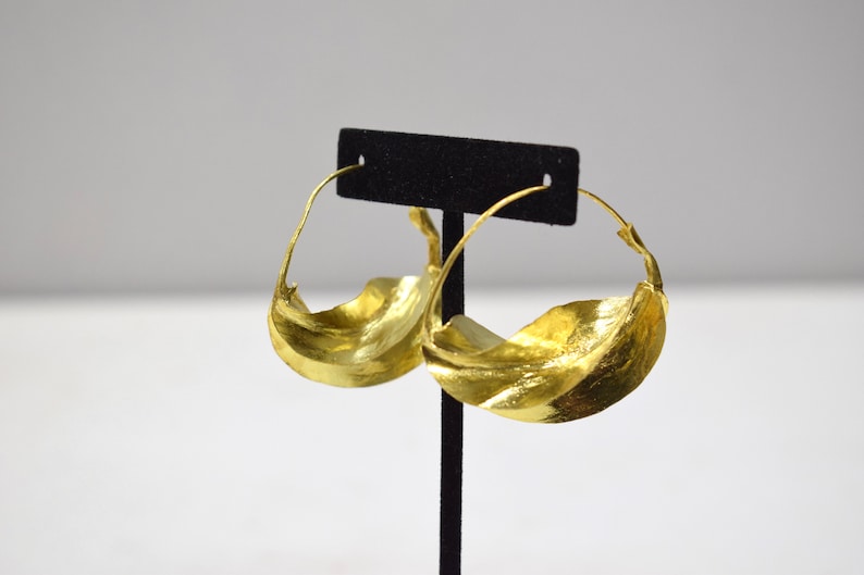 May include: A pair of gold hoop earrings with a wavy, organic design. The earrings are displayed on a black stand.
