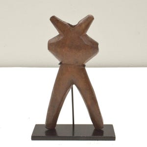 May include: A wooden sculpture of a stylized human figure with a triangular head and long legs. The sculpture is mounted on a black stand.