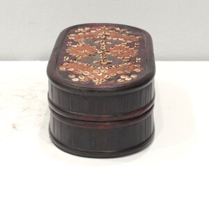 May include: A dark brown wooden box with a decorative pattern of small, light brown shells on the top. The box has a rounded oval shape and is made of wood.