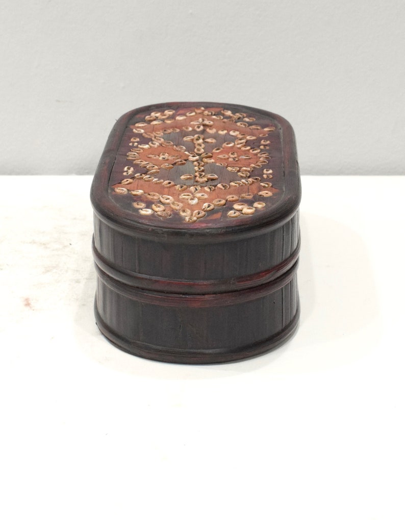 May include: A dark brown wooden box with a decorative pattern of small, light brown shells on the top. The box has a rounded oval shape and is made of wood.