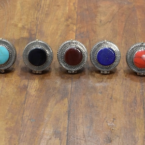 May include: Five round silver trinket boxes with different colored stones in the center. The stones are turquoise, black, red, blue, and red.