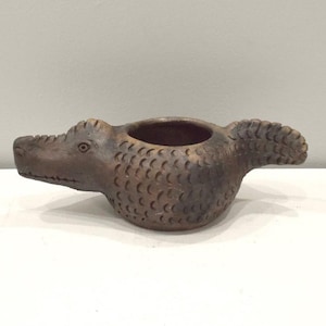 May include: A brown ceramic alligator-shaped planter with a textured surface. The planter has a small opening on the top for planting.