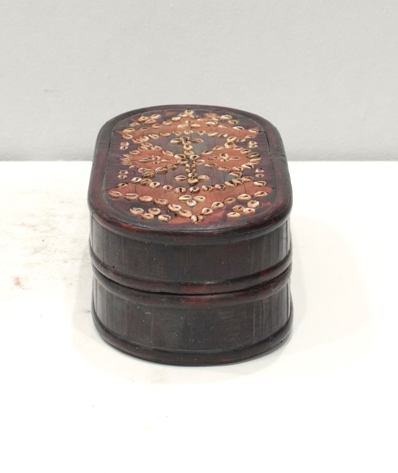 May include: A dark brown wooden box with an oval shape. The top of the box is decorated with a pattern of small, light brown shells.