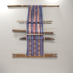 Pode incluir: Um tear de madeira com fios coloridos em tons de azul, roxo e vermelho. O tecido tecido apresenta um padrão geométrico. O tear está montado numa parede cinza claro, mostrando a arte têxtil.