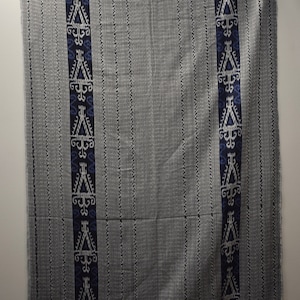 Op de afbeelding: Een grijze en witte geweven stof met een blauw en wit geometrisch patroon. De stof heeft een gefranst randje.