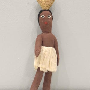 Könnte beinhalten: Handgefertigte Puppe mit brauner Haut, schwarzem Haar und rotem Mund. Die Puppe trägt einen cremefarbenen Rock und einen geflochtenen Korb auf dem Kopf. Die Arme der Puppe sind am Körper angenäht, und die Beine sind leicht angewinkelt.