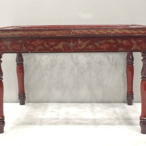 Puede incluir: Una mesa rectangular de madera roja con diseños florales y de vid dorados. La mesa tiene cuatro patas torneadas en una combinación de colores rojo y marrón a juego. La mesa es un mueble decorativo.