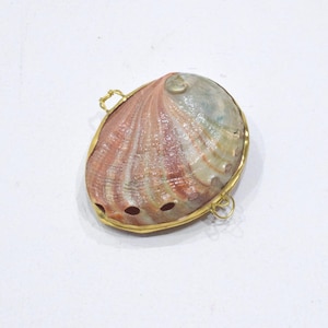 May include: An abalone shell pendant featuring a natural iridescent surface with pink, green, and cream tones. The shell is encased in a thin gold-toned metal frame with two small circular loops attached to the sides for hanging or attachment.