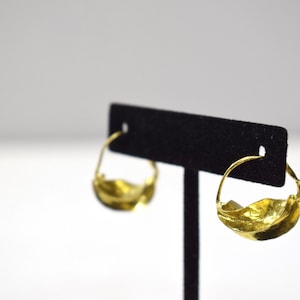 May include: A pair of gold hoop earrings with a wavy, organic design. The earrings are displayed on a black velvet stand.