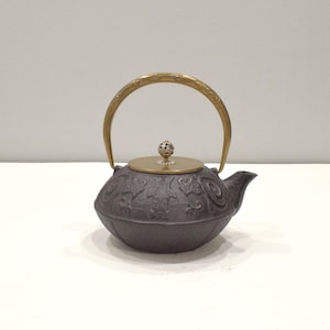 Puede incluir: Una tetera de hierro fundido de color gris oscuro con un asa y una tapa doradas. La tetera tiene un diseño floral ornamentado y un pequeño pico. El asa se arquea sobre la parte superior y la tapa está rematada con un pomo decorativo.