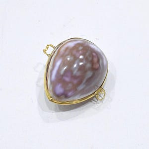 May include: A teardrop-shaped pendant featuring a polished, mottled brown and purple seashell encased in a thin gold-toned metal frame. The setting includes two small, delicate gold-toned wire loops attached to the sides for suspension.