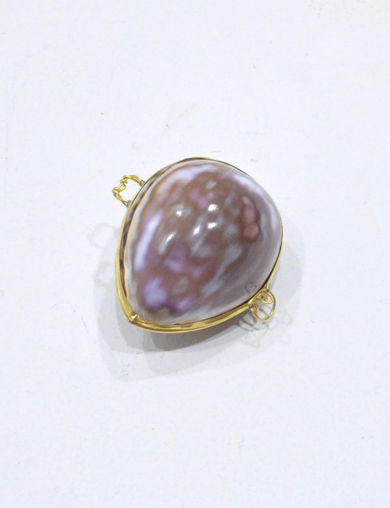 May include: A teardrop-shaped pendant featuring a polished, mottled brown and purple seashell encased in a thin gold-toned metal frame. The setting includes two small, delicate gold-toned wire loops attached to the sides for suspension.
