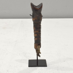 May include: A wooden carving of a stylized human figure with a long, thin body and a rounded head. The figure is painted in shades of brown and black and has a rope tied around its waist. The figure is mounted on a black stand.