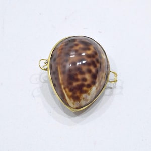May include: A teardrop-shaped cowrie shell pendant featuring a glossy brown and tan spotted pattern. The shell is encased in a thin gold-toned metal frame with small loops on both sides, designed for attachment to a chain or cord for jewelry.