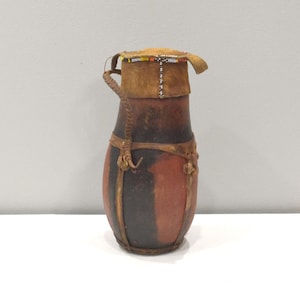Puede incluir: Una pequeña calabaza de cuero marrón con una tapa de cuentas y un asa de cuero. La calabaza está decorada con rayas negras y marrones.