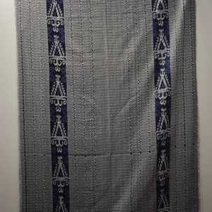 Op de afbeelding: Een grijze en witte geweven stof met een blauw en wit geometrisch patroon. De stof heeft een gefranst randje.