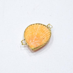 May include: A scallop shell pendant featuring a natural orange and white ribbed texture, encased in a thin gold-toned metal frame with two small wire loops attached to the sides for hanging or connecting to a chain or jewelry component.