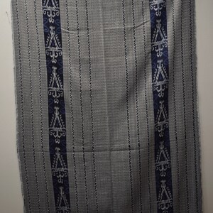 Op de afbeelding: Een grijze en witte geweven stof met blauwe en witte geometrische patronen. De stof heeft een gefranst randje.